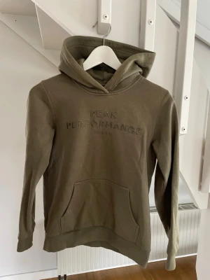 Olivgrön hoodie från Peak Performance - Snygg olivgrön hoodie från Peak Performance med broderad logga på bröstet. Klassisk känguruficka framtill och ribbade muddar vid ärmar och nederkant. Tillverkad i mjuk bomullsblandning och har en stor huva för extra komfort.