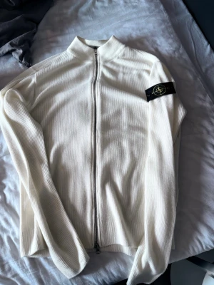 Vit stickad ziptröja från Stone Island - Säljer en stilren vit stickad ziptröja från Stone Island med ribbad struktur och hög krage. Tröjan har hel dragkedja framtill och den klassiska svarta Stone Island-loggan på vänster ärm. Perfekt för lager-på-lager och enkel att matcha.
