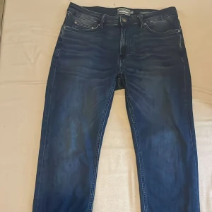 Mörkblå raka jeans - Säljer ett par mörkblå jeans med rak passform och klassisk femficksdesign. Jeansen har en stilren svart patch bak med texten 'SINCE 1969'. Materialet är denim i bomull och de har subtila slitningar för en avslappnad look.