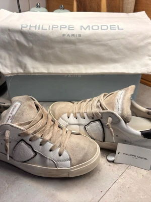 Philippe Model vita sneakers - Snygga vita sneakers från Philippe Model med beige mockadetaljer och vita läderpaneler. Skorna har en låg silhuett, beigea skosnören och den ikoniska sköldloggan på sidan. Perfekta för en trendig och avslappnad stil.