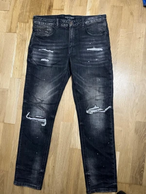Svarta slitna jeans med hål - Säljer ett par svarta jeans från Supply & Demand med slitningar och hål på knäna. Jeansen har en skinny fit, fem fickor och coola detaljer som blekta partier och små färgstänk. Perfekta för en trendig streetwear-look.