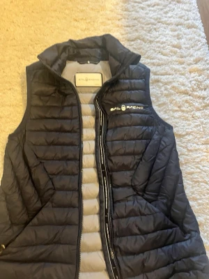 Svart dunväst från Sail Racing - Snygg svart dunväst från Sail Racing med quiltad design och hög krage. Västen har dragkedja framtill och logga broderad på bröstet. Insidan är ljusgrå och materialet är lätt och värmande, perfekt för lager-på-lager-stil.