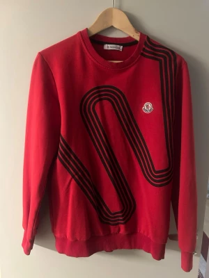 Röd stickad tröja från Moncler - Röd stickad tröja från Moncler med svart grafiskt mönster på framsidan och Moncler-logga på bröstet. Tröjan har rund halsringning, långa ärmar och ribbade muddar. Perfekt för dig som gillar streetwear och vill sticka ut.