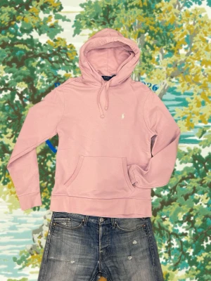 Ljusrosa hoodie från Polo Ralph Lauren - Säljer en ljusrosa hoodie från Polo Ralph Lauren med klassisk huva och dragsko. Tröjan har en känguruficka framtill och den ikoniska vita logon broderad på bröstet. Tillverkad i mjukt bomullsmaterial som är perfekt för chill dagar.