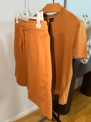 Orange set från BOSS med t-shirt & shorts - Snyggt orange set från BOSS med matchande t-shirt och shorts. Setet har en avslappnad passform och är gjort i mjukt bomullsmaterial. Både t-shirten och shortsen har diskret BOSS-logga. Perfekt för en chill och trendig look. Storlek L passar som M