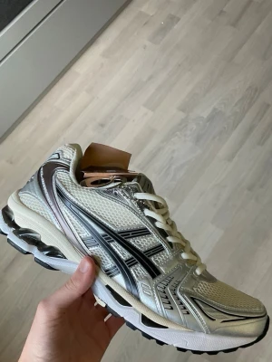 Asics beige och silver sneakers - Snygga sneakers från Asics i beige och silver med svarta detaljer. Ovandelen är i mesh och syntetmaterial för bra andningsförmåga och komfort. Klassisk Asics-logga på sidan och vita skosnören. Sulan är vit med svarta inslag och ger bra stöd.