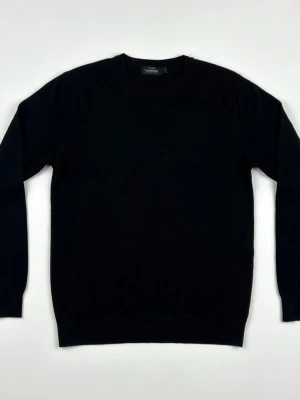 Svart stickad tröja från Dressmann - Klassisk svart stickad tröja från Dressmann i mjuk bomull. Tröjan har rund halsringning, långa ärmar och ribbade muddar vid ärmslut och nederkant. Perfekt basplagg för en clean och stilren look. Nypris runt 400kr