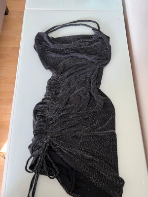 Svart glittrig kort klänning - Säljer en svart glittrig kort klänning med smala axelband och dragsko i sidan för snygg rynkeffekt. Klänningen har en figurnära passform och är perfekt för dig som vill sticka ut. Materialet är mjukt och stretchigt syntet. Använd en gång.
