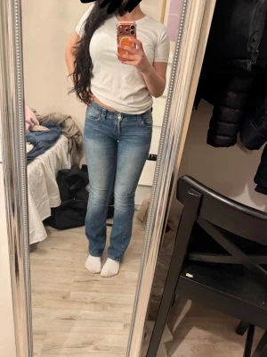 Blå bootcut jeans med fickdetaljer - Säljer ett par snygga blå bootcut jeans med klassisk femficksdesign och dekorativa ficklock bak. Jeansen har en lätt tvättad look och sitter tajt över låren för att sedan bli vidare nedtill. Perfekta till sneakers eller boots!