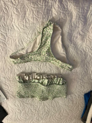Grön blommig bandeau bikini - Söt bandeau bikini med grönt blommönster och volangdetalj upptill. Överdelen är smockad och axelbandslös, medan underdelen har klassisk bikini-trosa form. Perfekt för lata dagar på stranden.