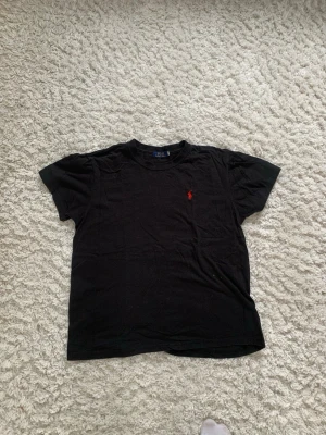 Svart t-shirt från Polo Ralph Lauren - Klassisk svart t-shirt från Polo Ralph Lauren med röd broderad logga på bröstet. T-shirten har rund hals och korta ärmar, perfekt basic-plagg till garderoben. Tillverkad i mjuk bomull för skön känsla.