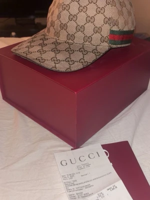 Beige keps från Gucci med GG-mönster - Säljer en beige keps från Gucci med det klassiska GG-monogrammönstret över hela kepsen. På sidan finns en grön och röd rand som ger en extra lyxig touch. Kepsen är tillverkad i canvasmaterial och har en böjd skärm. Perfekt för dig som gillar exklusiva accessoarer.