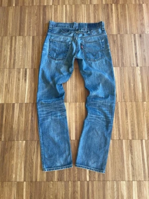 Levi's 501 blå jeans straight fit - Klassiska Levi's 501 jeans i blå tvätt med raka ben och normal passform. Jeansen har fem fickor, knappgylf och den ikoniska röda Levi's-taggen på bakfickan. Tillverkade i slitstarkt bomullsdenim med snygga slitningar och kontrastsömmar.