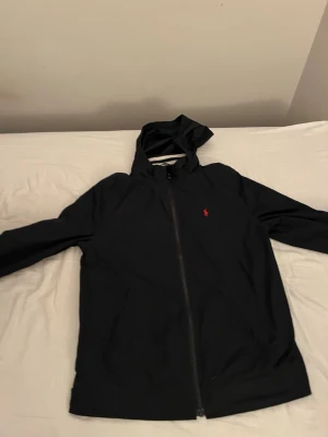 Marinblå vindjacka från Polo Ralph Lauren - Snygg marinblå vindjacka från Polo Ralph Lauren med huva och dragkedja framtill. Jackan har det klassiska röda Polo-logotypen broderad på bröstet. Tillverkad i lätt och vindtätt material, perfekt för blåsiga dagar.