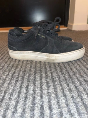 Axel Arigato sneakers - Snygga svarta sneakers från Axel Arigato i mocka med vit sula och diskret logga i guld på sidan. Klassisk låg modell med snörning och stilren design som passar till det mesta.