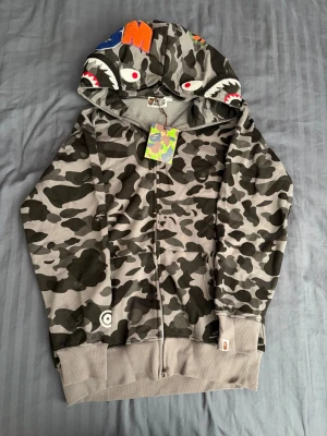 BAPE 1st Camo Shark Full Zip Hoodie - Säljer en helt ny grå camo zip hoodie från BAPE med ikonisk hajdesign på huvan och färgglada bokstäver. Hoodien har hel dragkedja, ribbade muddar och klassisk streetwear-look. Perfekt för dig som vill sticka ut med en unik och eftertraktad hoodie. L/M storlek