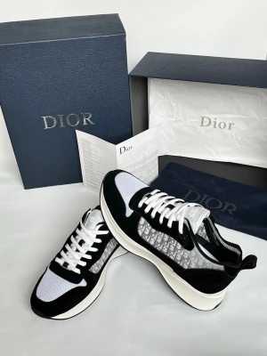 Dior sneakers svart/vit mesh - Snygga Dior sneakers i svart och vitt med meshpaneler och klassiskt Dior-mönster på sidan. Skorna har vita snören, svart mockadetalj och tjock vit sula för en modern look. Perfekta för dig som vill ha något exklusivt och trendigt.