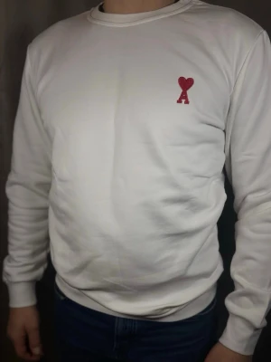 Vit sweatshirt från Ami Paris - Vit sweatshirt från Ami Paris med röd broderad logga med hjärta och bokstaven A på bröstet. Tröjan har rund halsringning och långa ärmar med ribbade muddar. Perfekt för en clean och stilren look.