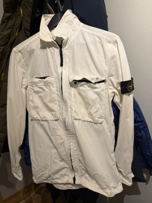 Vit overshirt från Stone Island - Snygg vit overshirt från Stone Island med klassisk patch på vänster ärm. Jackan har två stora bröstfickor med dragkedja och hel dragkedja framtill. Tillverkad i lätt bomullsmaterial och har en rak passform med hög krage. Perfekt för lager-på-lager och streetwear.