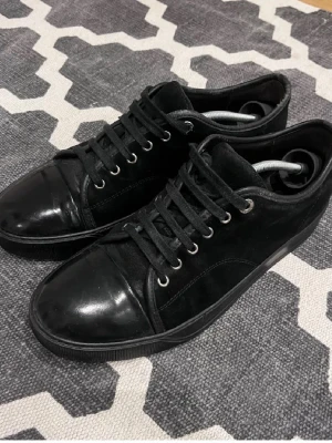 Svarta sneakers i mocka och läder - Stilrena svarta sneakers med ovandel i mocka och detaljer i blankt läder. Skorna har svarta skosnören, metallfärgade öljetter och en klassisk låg profil. Passar perfekt till streetwear och casual outfits.