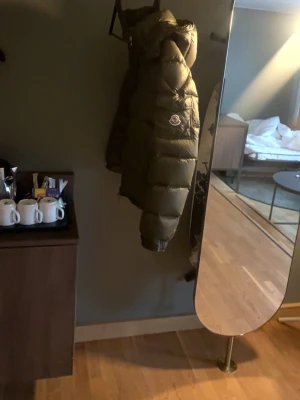 Olivgrön dunjacka från Moncler - Säljer en olivgrön dunjacka från Moncler med klassisk puffer-design och logotyp på bröstet. Jackan har hög krage och quiltade paneler som ger en riktigt schysst look. Perfekt för kalla dagar när du vill hålla värmen och ändå se snygg ut.