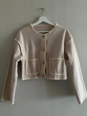 Ljusbeige kofta med dekorativa kanter - Söt ljusbeige kofta med dekorativa virkade kanter i vitt, rund halsringning och fyra knappar framtill. Två stora fyrkantiga fickor framtill ger en trendig look. Koftan är croppad och har långa ärmar, perfekt för lager på lager.