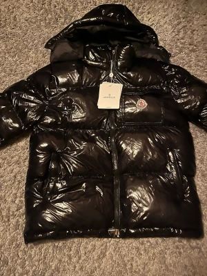 Svart glansig dunjacka från Moncler - Säljer en svart, glansig dunjacka från Moncler med huva och dragkedja framtill. Jackan har en bröstficka med Moncler-logga och stora, puffiga sektioner som ger en riktigt trendig look. Perfekt för kalla vinterdagar och riktigt snygg streetstil.