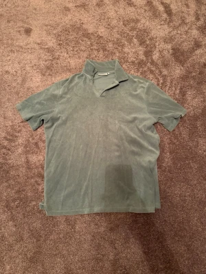 Grön kortärmad polotopp från Dobber - Säljer en grön kortärmad polotopp från Dobber i mjuk frotté. Toppen har en avslappnad krage och rak passform, perfekt för dig som gillar en chill och enkel stil. Passar till både jeans och shorts. Storlek M