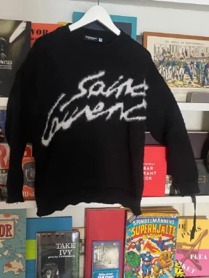 Svart stickad Saint Laurent-tröja - Säljer en svart stickad tröja från Saint Laurent med vit, stor logotyp i graffiti-stil på framsidan. Tröjan har rund hals, långa ärmar och ribbade muddar. Perfekt för dig som gillar streetwear och exklusiva märken.