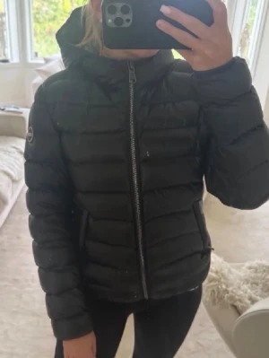 Svart pufferjacka från colmar - Säljer en svart pufferjacka från colmar med huva och dragkedja framtill. Jackan har quiltad design och en patch på ärmen. Perfekt för kyliga dagar och riktigt skön att bära. Passar dig som gillar en clean och sportig look. Svart på utsidan och grå på insidan. Ena fickan är trasig och går inte att stänga och kan förekomma hål som jag inte har sett då den är köpt begagnad. Lånad bild jackan är inte glansig