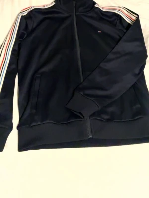 Marinblå ziptröja Tommy Hilfiger - Snygg marinblå ziptröja från Tommy Hilfiger med klassiska ränder i rött, vitt och blått längs ärmarna. Tröjan har hög krage, hel dragkedja och två fickor framtill. Liten logga på bröstet och ribbade muddar vid ärmslut och nederkant.