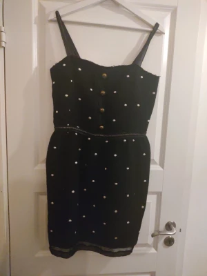 Svart miniklänning med sydda vita prickar. Marc Jacobs  - Snygg svart miniklänning med vita prickar och smala axelband. Klänningen har dekorativa knappar framtill och dragkedja i ryggen. Modellen är figurnära med markerad midja. Perfekt för dig som gillar en lekfull men stilren look.