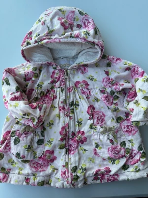 Blommig jacka med huva från Baby - Supersöt jacka i vit manchester med rosa rosor och gröna blad. Jackan har dragkedja framtill, två fickor med rosett och en mysig huva. Perfekt för dig som gillar romantiska och gulliga plagg.tvättpåverkan finns