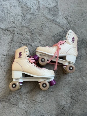 Beige retro rullskridskor Story - Snygga beige rullskridskor från Story med rutmönster och rosa detaljer. Klassisk retrostil med högt skaft, rosa snören och lila bromsklossar. Hjul i beige och vitt, perfekt för dig som vill sticka ut på rinken eller gatan. Sparsamt använda, skavanker finns(se bilder). Priset kan diskuteras!