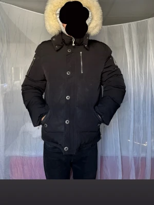 Svart parkas med äkta päls - Moose Knuckles - Svart Moose Knuckles jacka med stor huva och vit päls. Jackan har dragkedja och knappar framtill, bröstficka med dragkedja och fickor på sidorna. Ärmficka med dragkedja för extra stil. Perfekt för kalla dagar och riktigt snygg. Välanvänd men i fint skick. Går att lämna in på kemtvätt för att få bort små fläckar, se bilderna. Storlek 2XL men passar även L.