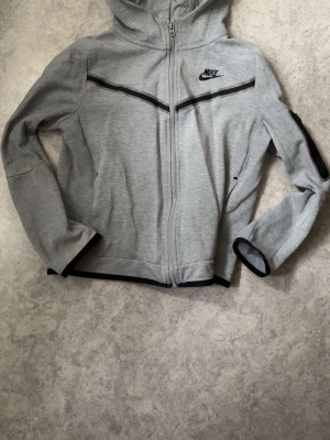 Grå nike tech fleece med dragkedja - Snygg grå hoodie från Nike med svart logga på bröstet och svarta detaljer längs dragkedjan och ärmsluten. Hoodien har hel dragkedja, huva och en ficka på ärmen. Tillverkad i mjukt sweatshirtmaterial, perfekt för en sportig look.