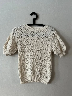 Vit stickad topp med puffärm - Söt vit stickad topp med korta puffärmar och vågigt hålmönster. Rund halsringning och ribbade muddar vid ärmslut och nederkant. 