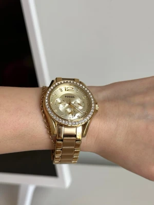 Fossil Guldig klocka med stenar - Snygg guldig klocka från Fossil med länkarmband och glittrande stenar runt urtavlan. Klockan har flera små urtavlor och en stilren design som verkligen sticker ut. Perfekt accessoar för dig som gillar lite extra bling.