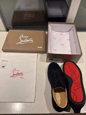  svarta sneakers - Säljer ett par svarta sneakers från med ikoniska röda sulor och svarta nitar på tån. Skorna har en låg profil och är tillverkade i mocka och läder. Kommer med originalkartong, dustbag och extra nitar. Perfekta för dig som vill sticka ut.