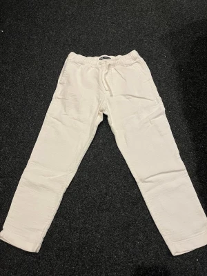 Vita linnebyxor från Zara - Säljer ett par vita linnebyxor från Zara med avslappnad passform och resår i midjan samt dragsko. Byxorna har sidofickor och är tillverkade i linnematerial blandat med bomull därmed är dem lite tjockare. Endast anävnda 2 gånger.