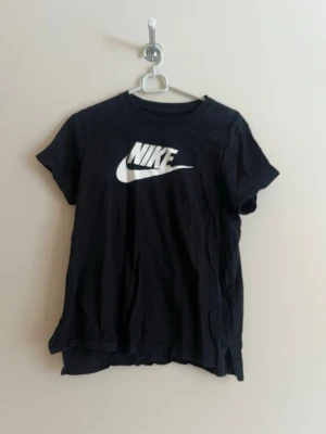 Svart Nike t-shirt med logga - Snygg svart t-shirt från Nike med klassisk vit logga på bröstet. T-shirten har rund halsringning och korta ärmar. Tillverkad i mjuk bomull som känns skön mot huden. Perfekt för en sportig och avslappnad stil.