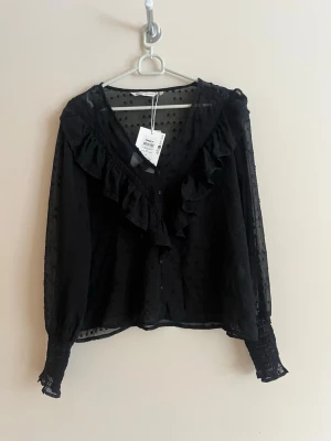 Svart volangblus från Only - Trendig svart blus från Only med volangdetaljer längs V-ringningen och knappar framtill. Blusen är tillverkad i ett transparent, prickigt mesh-tyg och har långa ärmar med smock vid ärmsluten. Perfekt för en cool och stilsäker look.