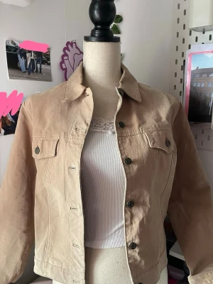 Suede jacka med klassisk krage - Snygg beige suede jacka med klassisk krage och knappstängning framtill. Två bröstfickor med lock och knappar ger en tidlös look. Jackan har raka ärmar och en avslappnad passform, perfekt att slänga över en topp för en cool vardagsstil. Äkta mocka 