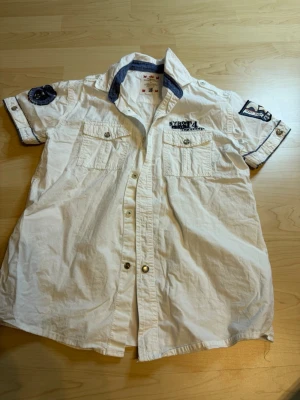 Vit skjorta från KappAhl med detaljer - Vit kortärmad skjorta från KappAhl med blå detaljer på kragen och coola tygmärken på ärmarna. Två bröstfickor med knappar och tryck på vänster sida. Perfekt för en avslappnad men snygg look.