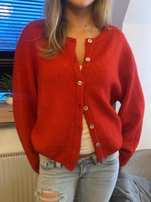 Röd oversized kofta med knappar - Mysig röd kofta i oversized modell med rund halsringning och silvriga knappar framtill. Koftan har långa ärmar och ribbade muddar vid ärmslut och nederkant. Perfekt att slänga över en t-shirt för en chill look.