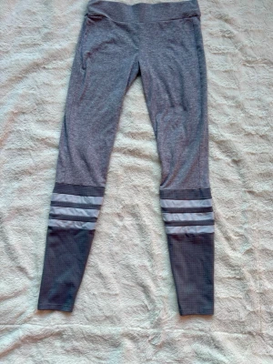 Grå leggings med reflexränder - Snygga grå leggings med bred midja och coola reflexränder runt vaderna. Nedre delen av benen har mörkare grå partier med diskret rutmönster. Perfekta för träning eller chill. Stretchigt material för skön passform.