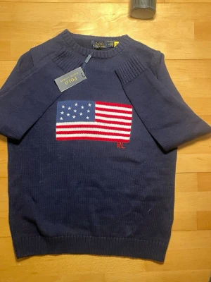 Mörkblå stickad tröja Polo Ralph Lauren - Mörkblå stickad tröja från Polo Ralph Lauren med amerikansk flagga i rött, vitt och blått på bröstet samt RL-broderi. Tröjan har rund halsringning och ribbade muddar vid ärmslut och nederkant. Tillverkad i mjuk bomull för en skön känsla. Såklart äkta ‼️‼️skriv vid minsta fundering 🤩med tags o allt