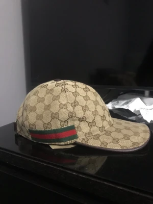 Gucci keps  - Säljer en beige keps från Gucci med det klassiska GG-monogrammet över hela kepsen. På sidan finns en grön och röd rand som ger den en ikonisk touch. Kepsen har böjd skärm och är tillverkad i canvasmaterial.
