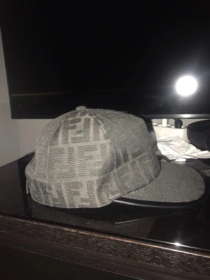 Fendi keps  - Säljer en grå keps från Fendi med det klassiska FF-logomönstret över hela kepsen. Kepsen har en rak skärm och är tillverkad i ett tygmaterial med detaljer i svart på skärmen. Perfekt för dig som gillar exklusiva accessoarer och streetwear. Knappt använd 