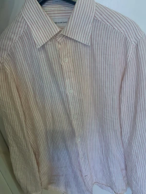 Randig skjorta i bomull från Blue Glory - Klassisk skjorta från Blue Glory i vit bomull med tunna röda ränder. Skjortan har lång ärm, knappar framtill och en avslappnad passform. Perfekt för dig som gillar en stilren och enkel look med lite extra detalj tack vare det randiga mönstret. Köpt på volt!